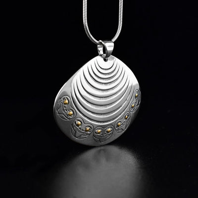 Sterling silver and 18K gold clam with frogs pendant created by Tahltan artist Grant Pauls. Design depicts a clam with frogs decorating the bottom. Eyes of the frogs are made from 18K gold and rest of piece is sterling silver. Measures 1.25" x 1.50". Chain not included.