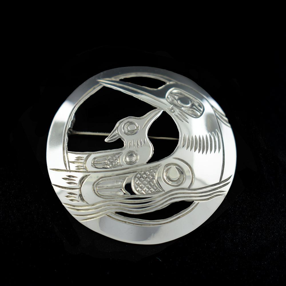 This sterling silver brooch is circle shaped and has cutouts and carvings that depict a mother and baby loon.