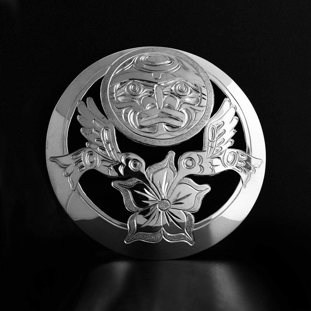 This Moon and Hummingbird pendant is made from sterling silver with a large circular shape. There are cutout designs and carvings on the surface of the pendant that depict Hummingbirds flying over a flower with the Moon in the back.