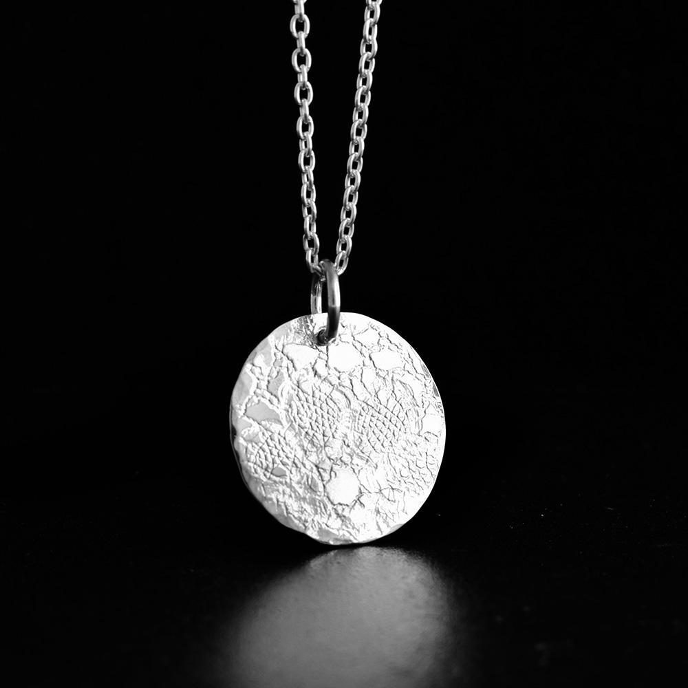 This lace pendant is made from sterling silver and is circle shaped and has lace designs on it. The designs have been achieved by embossing French lace into the surface of the piece.