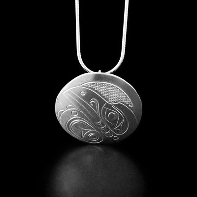 Sterling Silver Round Raven Pendant by Don Lancaster. The design depicts the profile of a raven's head facing the top left corner of the pendant. The raven has a long, narrow beak. The artist has carved a wing underneath the head of the raven with intricate designs representing the feathers. The "background" of the pendant has been neatly carved into a crisscross pattern to allow for the legend to stand out.