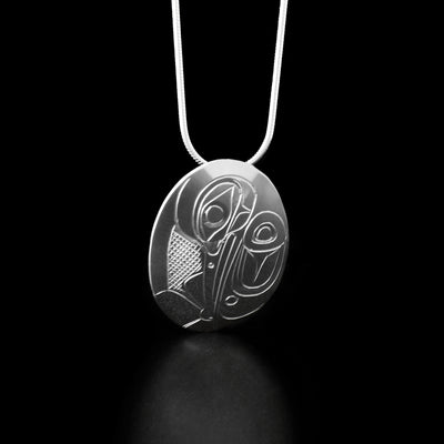 Sterling Silver Oval Hummingbird Pendant by Don Lancaster. The pendant is in the shape of an oval and the design depicts the profile of a hummingbird's head facing downwards. The hummingbird has a long, narrow beak and is drinking from a flower that the artist has hand-carved at the bottom of the pendant. The artist has also hand-carved a wing to the right of the hummingbird's head with intricate designs representing the feathers of the legend. The "background has been carved into a crisscross pattern.
