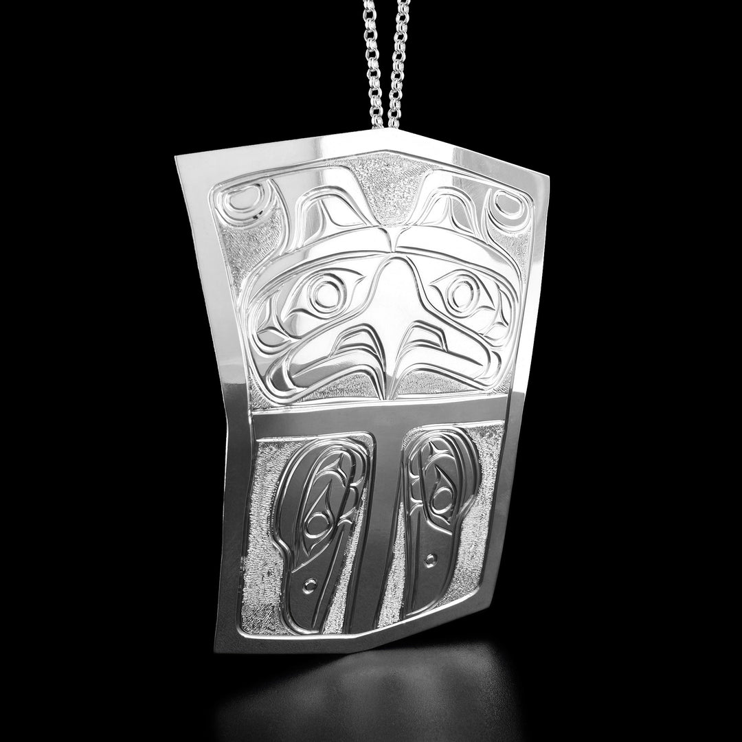 The top part of this shield pendant is carved into the face of a thunderbird looking straight ahead. The bottom part has two raven heads facing downward.