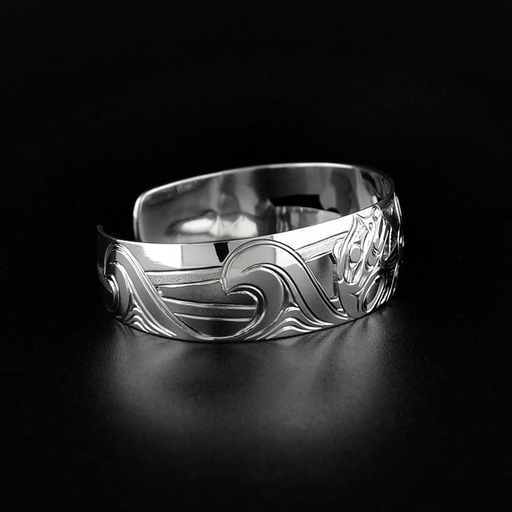 This Otter bracelet is made from sterling silver and has a single, wide band with a gap in the back. There is a depiction of the Otter laying on its back holding a sea urchin in the ocean.