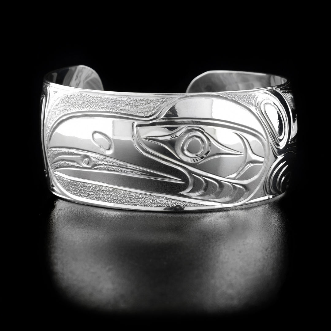 This Raven bracelet has a single, wide band with a gap in the back and is made form sterling silver. There is a large depiction of the raven that has been beautifully carved into the surface of the piece. There are textured designs made in the negative space around the legend.