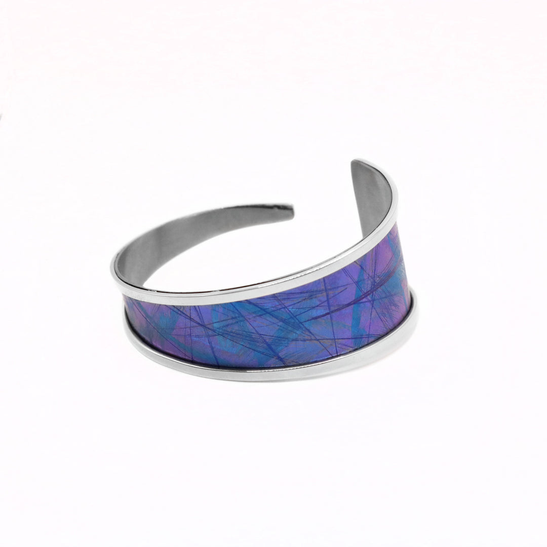 This titanium bracelet cuff has been made using sterling silver and anodized titanium. The piece is made up of a long, tapered titanium band with silver bands lining the sides of it and is curled up into a bangle with a small gap in the back. There is a dark blue and purple color with blue designs on the surface of it that are achieved by anodizing the metal.