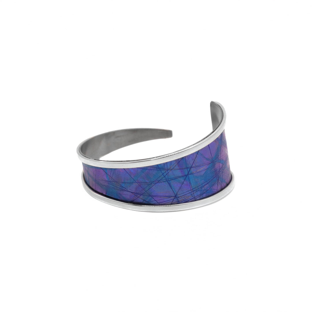 This titanium bracelet cuff has been made using sterling silver and anodized titanium. The piece is made up of a long, tapered titanium band with silver bands lining the sides of it and is curled up into a bangle with a small gap in the back. There is a dark blue and purple color with blue designs on the surface of it that are achieved by anodizing the metal.