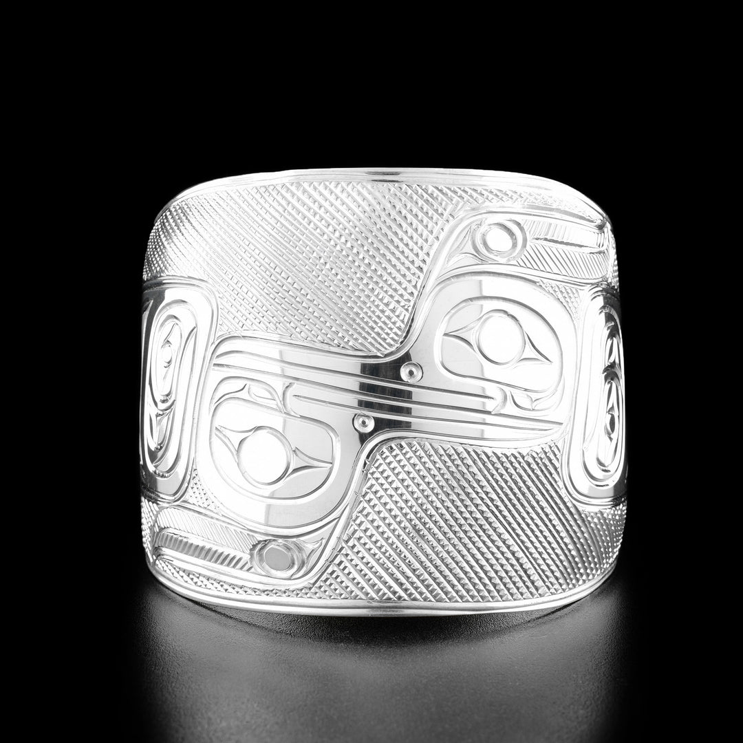 Sterling Silver Hummingbird Reflection Bracelet by Paddy Seaweed. The design depicts one hummingbird on the right side of the bracelet looking to the left. The second hummingbird is upside down on the left side of the bracelet looking to the right. The beaks are connected to make it appear as if the two hummingbirds were being reflected. The background has been carefully designed to allow for the image to stand out.