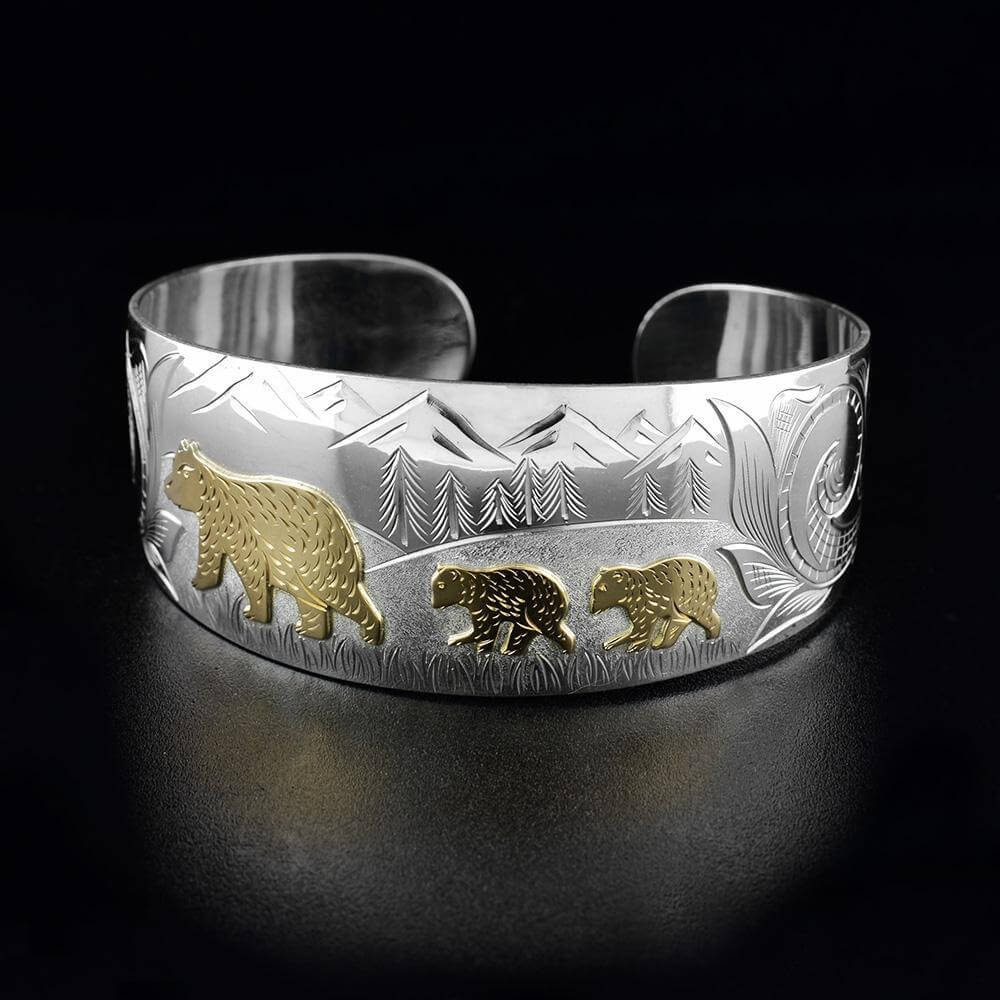 This bear family bracelet has a sterling silver, wide band with a gap in the back. There is a bear with two cubs done in 10K gold and there are bear designs and a mountain forest scene carved into the surface of the band.