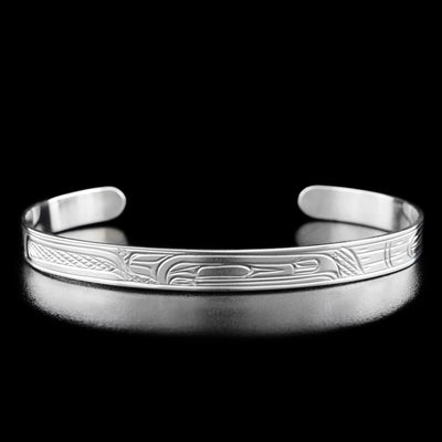 Sterling Silver 1/4" Raven Bracelet by Victoria Harper. In the center of the bracelet there is the profile of a raven's head facing towards the right. The raven has a large feather on the back of its head and a long, skinny beak. The artist has hand-carved feather-like designs to the right of the raven's head. To the left of the raven's head the artist has included more intricate designs representing the raven's wing and body.