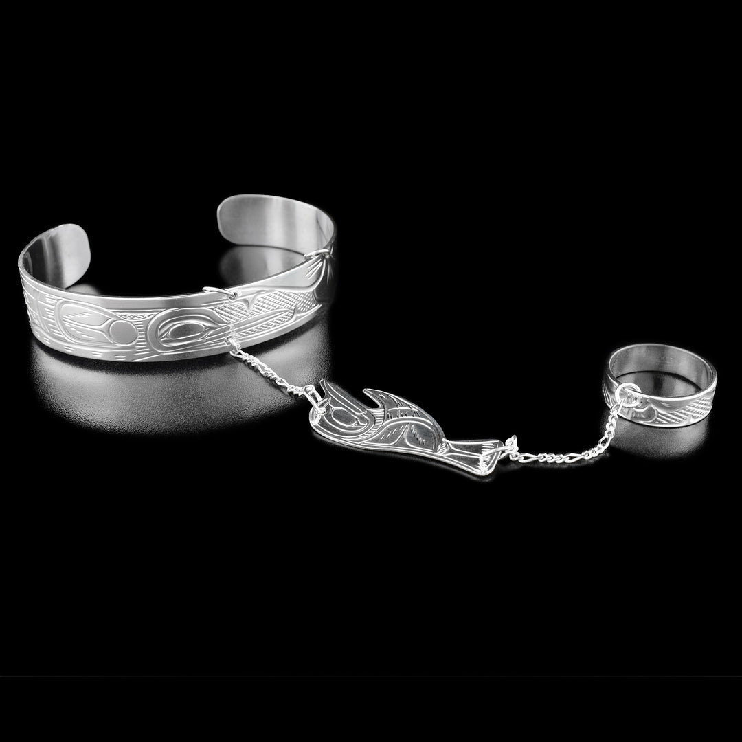 Hummingbird bracelet connected to a ring by a thin chain. In the center of the chain is a carved hummingbird accessory. The center of this hummingbird bracelet has the profile of a hummingbird mid-flight facing the left with an extended wing. To the right of the hummingbird are hand-carved flowers.