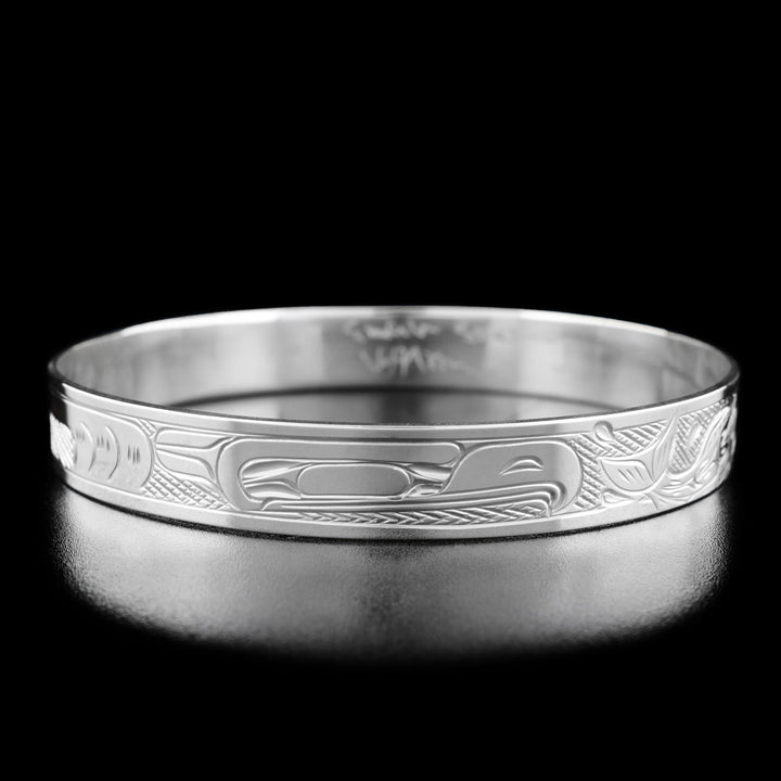 Sterling Silver 3/8" Eagle Chasing Salmon Bangle by Victoria Harper. In the center of the bracelet there is the profile of an eagle's head facing towards the right. To the right of the eagle's head there are two salmon swimming away with waves underneath them. To the left of the eagle the artist has hand-carved intricate designs to represent the eagle's feathers and body. The "background" of the bracelet has been hand-carved into a crisscross pattern to allow for the legends to stand out.