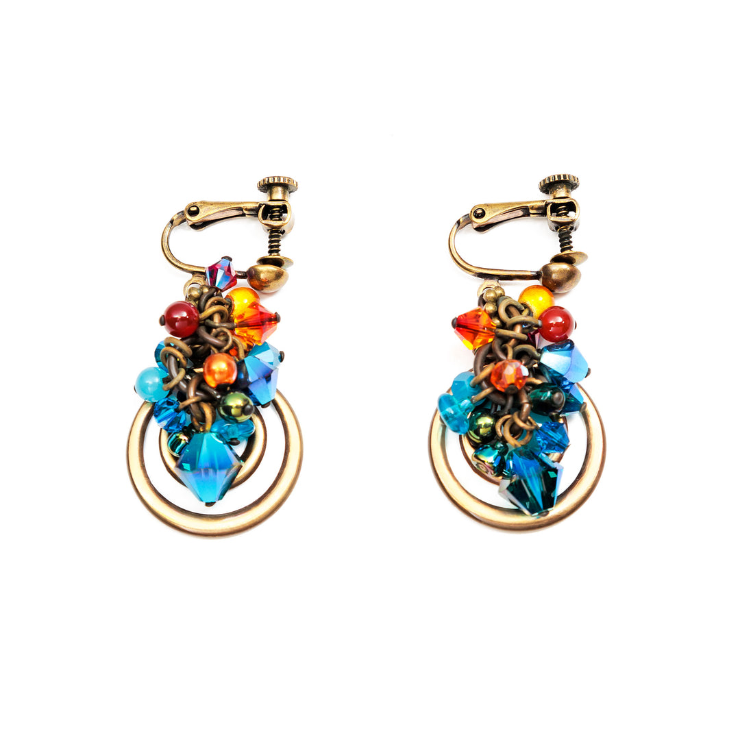These brilliant crystal cluster earrings are made from carnelian agate, Swarovski crystal, hand worked brass, and glass. There are blue, orange and red bead cluster with small beads made from agate and glass. The crystals are blue and make up the larger beads on the piece. Below the clusters are brass hoops, one being smaller and inside the other.