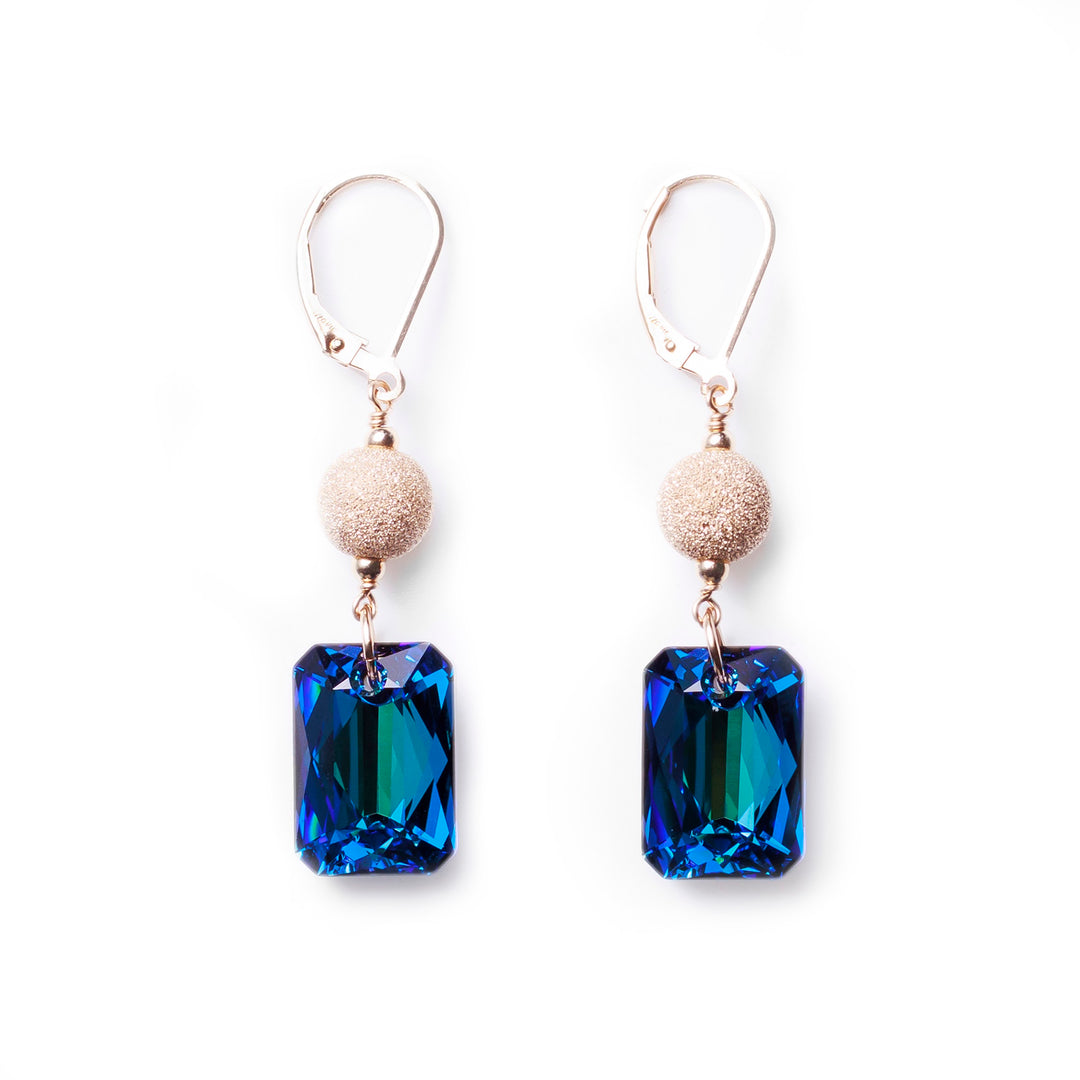 These Swarovski crystal drop earrings are made from Stardust beads, 14K Gold Filled wire, and bermuda blue Swarovski Crystals. The stardust beads are below the hooks and there are rectangular, emerald cut crystals. The hangs are dangling form the gold fill wire.