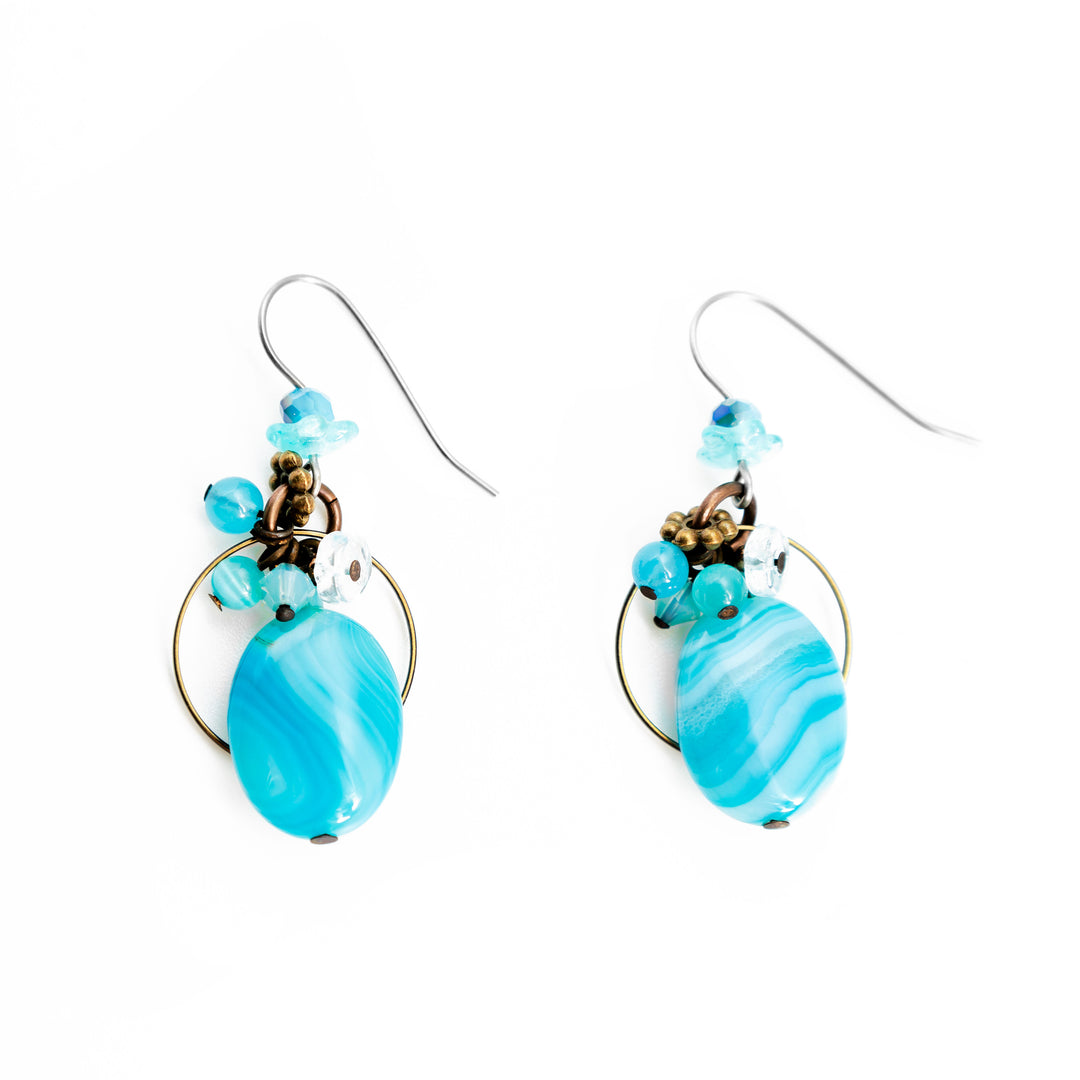 These aquamarine earrings have been made by hand worked brass, Austrian crystal, dyed agate, aquamarine, and glass. There is a cluster of beads below the hooks, a teardrop shaped aquamarine gem hung below them and a brass hoop behind it.
