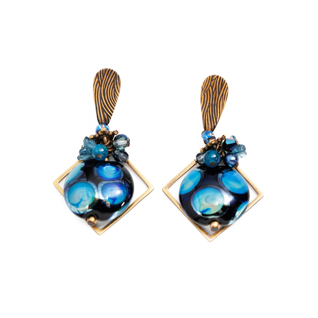 These dangling ball glass earrings are made using hand worked brass, B.C. artist glass, freshwater pearls, Austrian crystal, and glass. There is a cluster of crystal, glass and pearl beads below the hooks and below that is a circle shaped glass bead. The bead is dark blue and has interesting polka dot patterns on the surface of it. There is a off axis square hoop made of brass around the hangs.