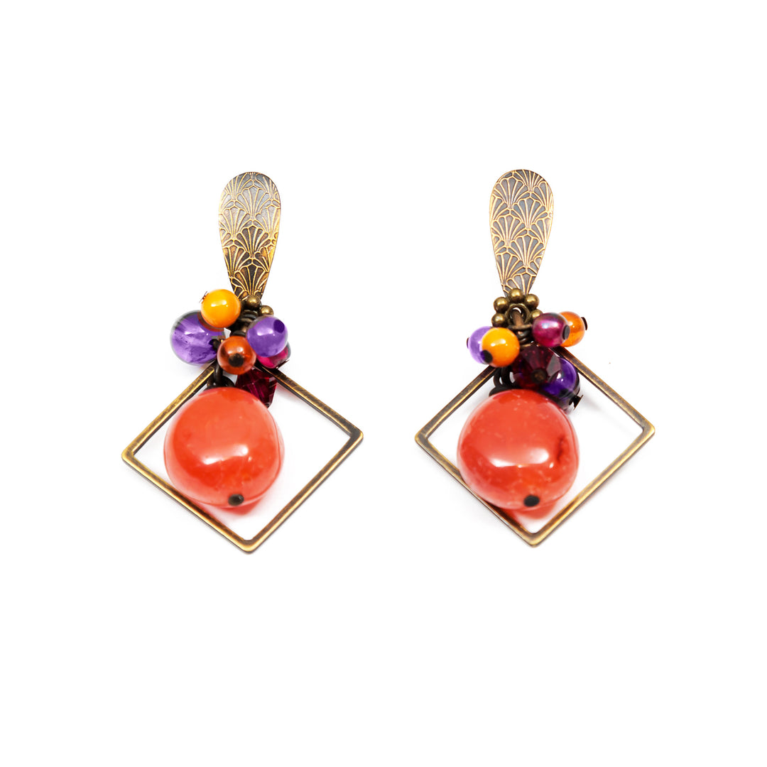 These funky agate earrings have been made using amber, carnelian agate, Austrian crystal, garnet, and mother of pearl. There is a cluster of beads made from the gems and crystal at the top of the hooks and below them is a large, polished and orange agate ball. There is a diamond shaped brass hoop framing the large bead. These pieces are studs and have dangling hangs to them.