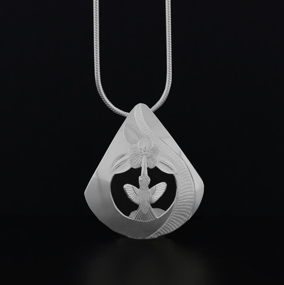 This Hummingbird and flower pendant is made from sterling silver and has a teardrop shaped pendant. There are cutout designs and carvings on it that depict the Hummingbird and a flower.