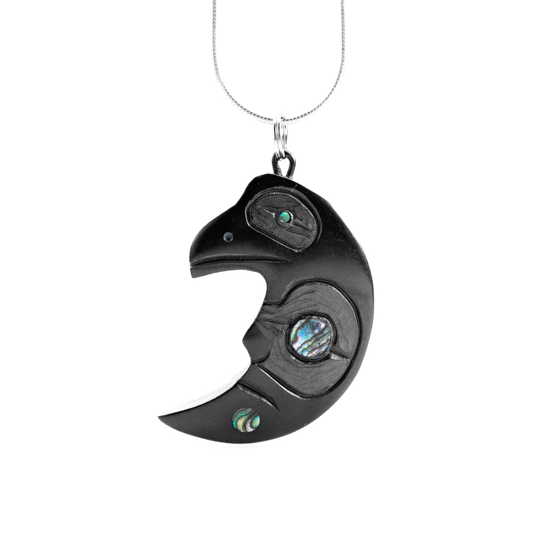 This Raven Moon pendant has been carved from argillite stone and has the shape of a crescent moon. At the top of the crescent, there is the head of the Raven with an abalone gem in its eye. The moon legend has a large piece of abalone as its eye and there are carvings across it depicting the legends. 