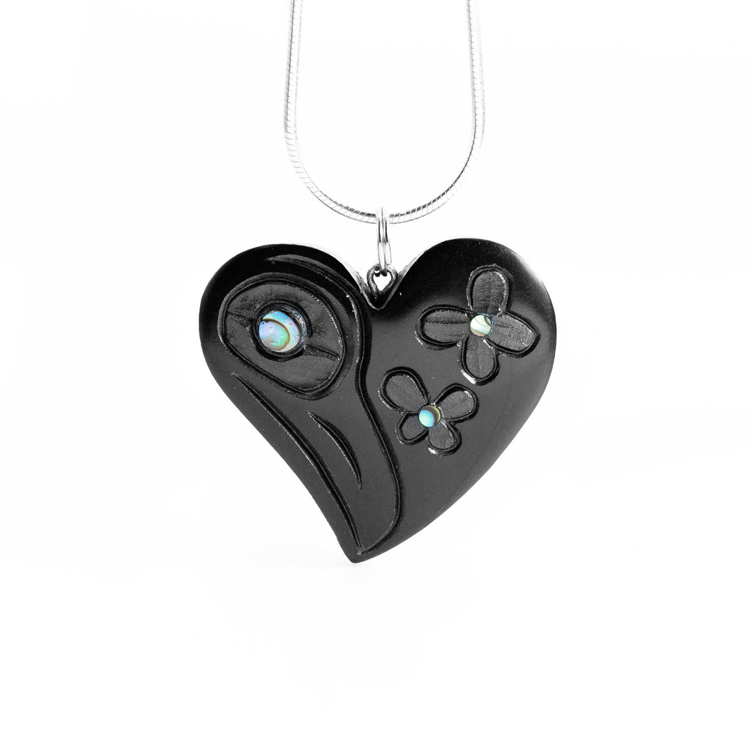 Argillite Hummingbird Heart Pendant by Amy Edgars. The pendant is in the shape of a heart. On the left side of the pendant the artist has hand-carved the profile of a hummingbird's head facing downwards. On the right side of the pendant the artist has hand-carved two small flowers. The artist has put abalone in the center of both flowers as well as in the hummingbird's eye.