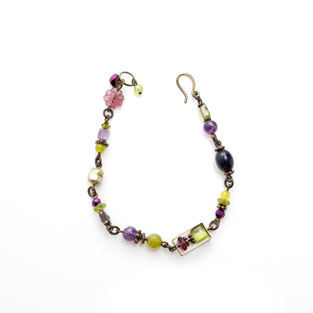 This pearl and crystal bracelet is made using hand worked brass, Austrian crystal, fresh water pearls, genuine amethyst, serpentine jade, peridot gems, and glass beads. The piece consists of beads with various shaped, sizes and materials (crystals, gems and glass) and the beads are connected using the brass. There are brace charms made around the beads. There is a large. pink flower bead featured on the piece.