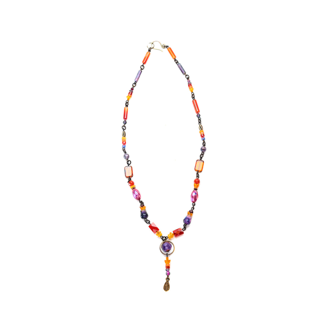 This beaded gemstone necklace is made from handworked brass, amethyst, glass, carnelian agate, dyed jade, aventurine, and carved amethyst tulips. The necklace features beads that have unique shapes, designs and materials. The beads are thin and and give the necklace a dainty and elegant look. There is a large amethyst gem at the bottom of the necklace and a small, beaded lariat that drops down below the piece.