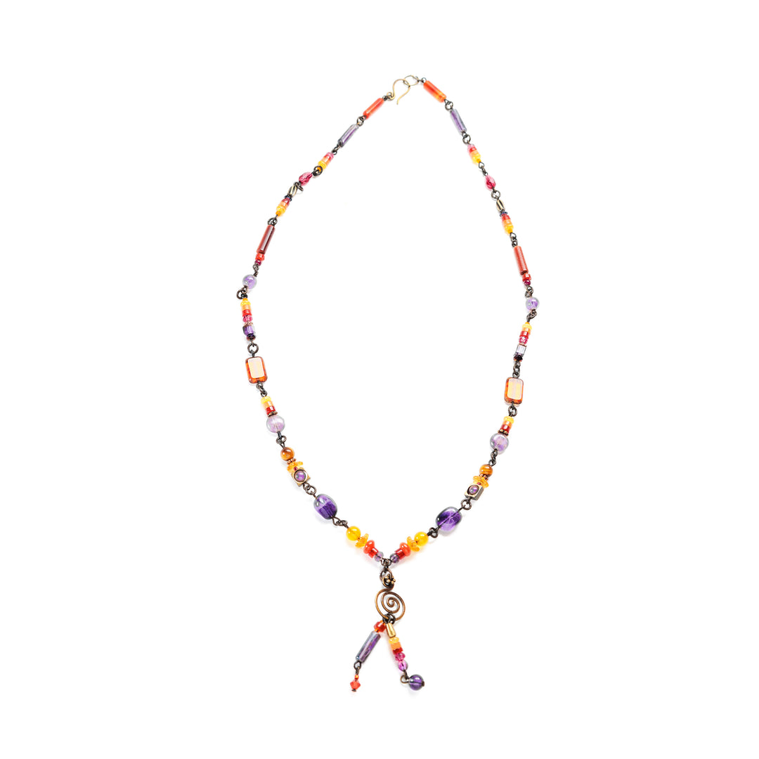 This beaded gemstone necklace is made from handworked brass, amethyst, glass, carnelian agate, dyed jade, aventurine, and carved amethyst tulips. The necklace features beads that have unique shapes, designs and materials. The beads are thin and and give the necklace a dainty and elegant look. There is a large amethyst gem at the bottom of the necklace and two small, beaded lariats that drops down below the piece.
