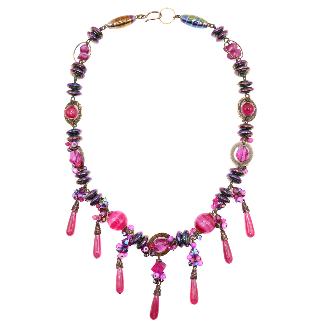 This gorgeous, beaded pink crystal necklace is made from Swarovski crystal, lampworked glass, handworked brass, agate, glass, and dyed calcite. The beads have unique designs, shapes and materials and give the piece a chunky look. There are long teardrop shaped gems dangling down from the bottom section of the piece and brass charms around the beads.