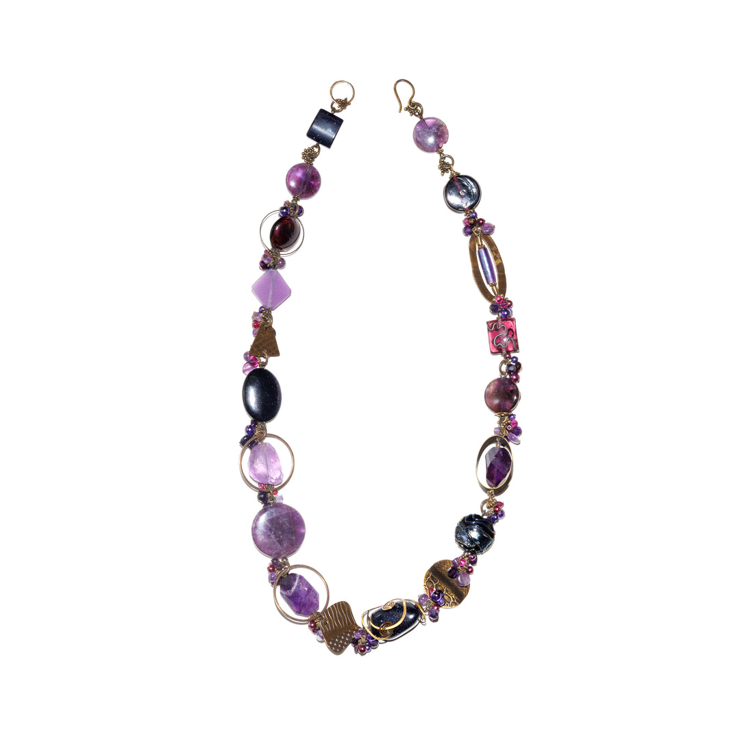 This funky amethyst necklace has chunky beads made from various designs, sizes and material (amethyst, brass, facetted glass, pressed glass, pearls, and fluorite). There are hand worked brass charms made around the piece and some frame the beads.