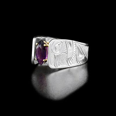 Solitaire Hummingbird Ring with Amethyst by Fred Myra. The artist has hand-carved the profile of two hummingbird bodies on either side of the amethyst he has set in the center of the ring. The background of the ring has been hand-carved into a neat crisscross pattern to allow for the hummingbirds to stand out. The amethyst has been set using a 14k gold claw setting. The ring tapers down at the back.