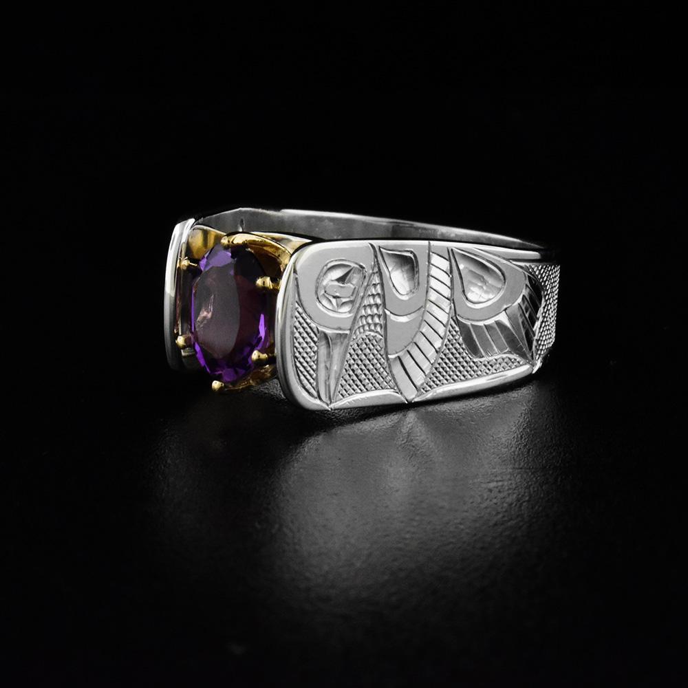 This cathedral ring has a wide, sterling silver band with a large, oval amethyst gem in the center. There is a depiction of the Hummingbird carved into both sides of the band in a side profile. The head is facing upwards towards the gem with its wing and tail below it. The bands extend above the gem in a cathedral fashion and the gem is held in 14K gold claws.