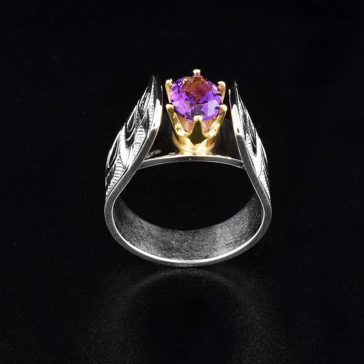 This cathedral ring has a wide, sterling silver band with a large, oval amethyst gem in the center. There is a depiction of the Hummingbird carved into both sides of the band in a side profile. The head is facing upwards towards the gem with its wing and tail below it. The bands extend above the gem in a cathedral fashion and the gem is held in 14K gold claws.