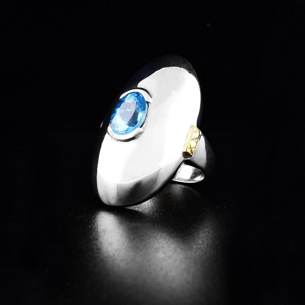 This blue topaz solitaire ring is made from sterling silver and has a large, raised oval shaped signet at the top of the ring. There is a large, topaz gemstone set into the signet and 14K gold highlights on the sides of the signet.