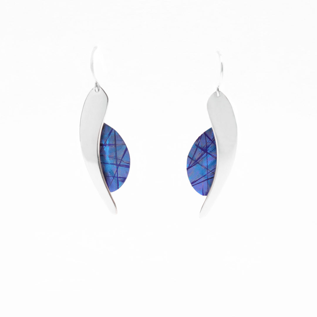 These titanium dangle earrings are made from sterling silver and anodized titanium. The hangs have a leaf shaped, titanium portion with a silver band running along the side of the piece. The titanium has a deep blue color and other blue colored designs on the surface of it that is achieved by anodizing the metal.