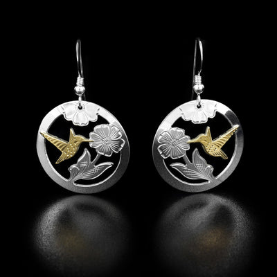 Each earring is a circular shape with a carved flower and a hummingbird made out of 10k yellow gold mid-flight drinking from it. The negative space has been cut out.