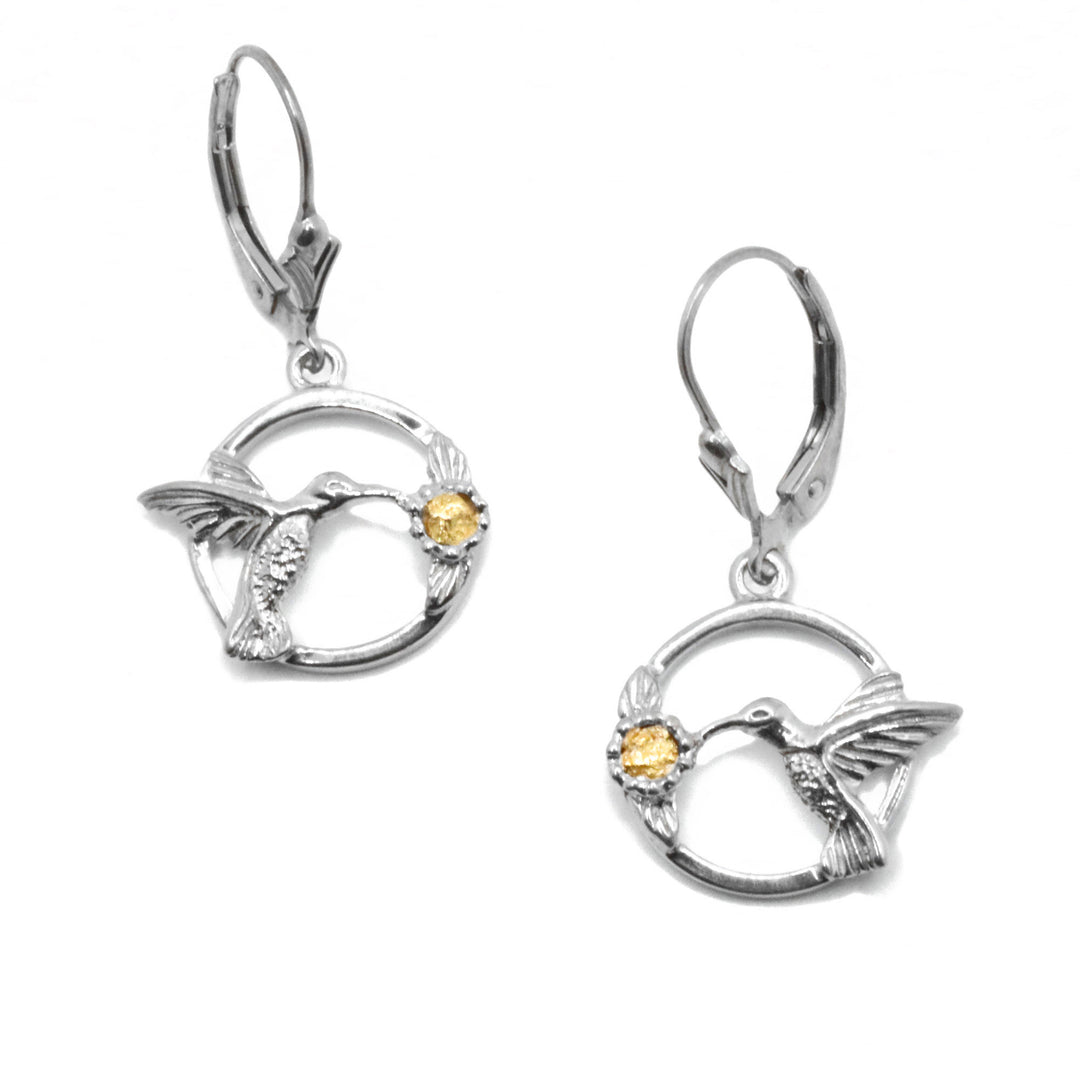 22K Gold Nugget Hummingbird Earrings by Tom Gregorczyk. Each earring has been handcrafted in a circular shape with a hummingbird mid-flight, drinking from a flower. The background has been cutout to create a 3D effect. The artist has included 22k gold nuggets in the center of both flowers. The rest of each earring is sterling silver.