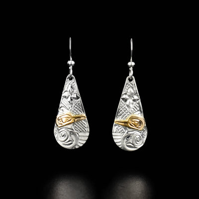 These hummingbird earrings are teardrop shaped. Each earring has the profile of a hummingbird with a spread wing and a 14k yellow gold head at the bottom. There is a flower carved above each hummingbird.