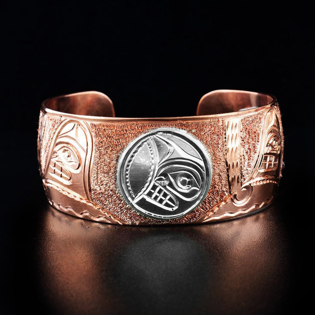 Sterling Silver and Copper 1" Orca Pod and Moon Bracelet by Paddy Seaweed. The design depicts 1/4th of the moon's face made from sterling silver in the middle of the bracelet. The remainder of the bracelet has various orcas swimming made from copper. The background has been neatly hand carved to create an eye-catching effect.