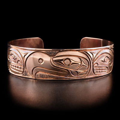 Copper 3/4" Thunderbird and Orca Bracelet by Paddy Seaweed. The design depicts the profile of a thunderbird's head looking to the left at the center of the bracelet. On both sides of the thunderbird are the profiles of an orca's head looking towards the thunderbird. Following the orca's heads are their bodies which have been delicately carved.