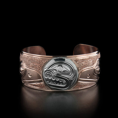 Sterling Silver and Copper Bear and Raven Bracelet by Paddy Seaweed. The designs depicts the profile of a bear's face, made out of sterling silver, in a circular shape in the center of the bracelet. On both sides of the bracelet are two profiles of ravens facing towards the bear made out of copper. The background has been delicately hand carved to allow for the legends to stand out.