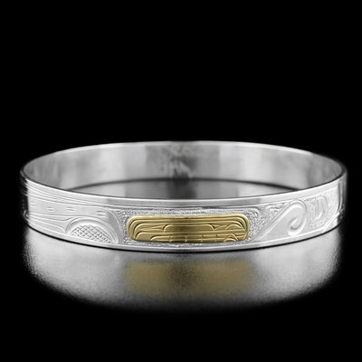 Sterling Silver and 14K Gold 3/8" Orca Bangle by Victoria Harper. In the center of the bracelet the artist has hand-carved the profile of an orca's head facing towards the right out of 14k gold. To the left of the orca's head is the continuation of the orca's body with fins and a tail made out of sterling silver. To the right of the orca's head the artist has hand-carved waves and the bodies of more orcas.