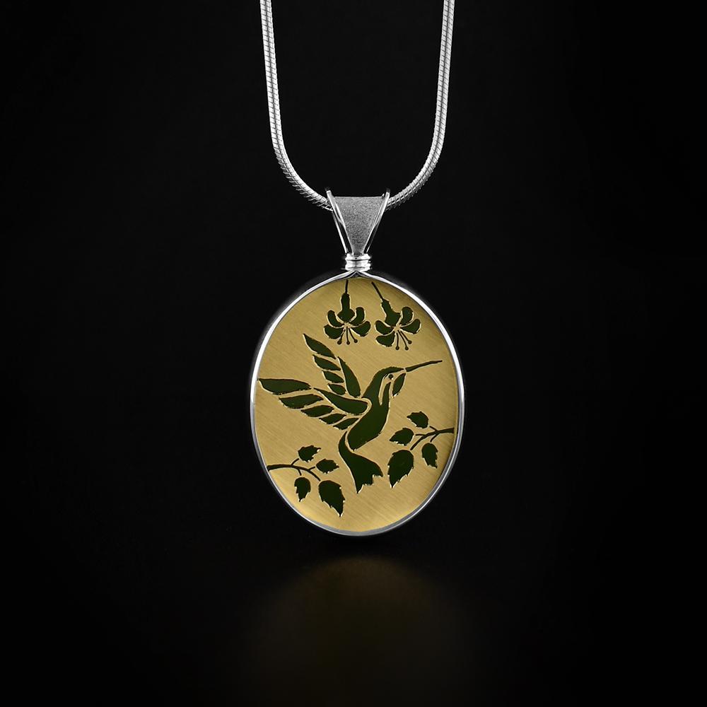 This Hummingbird pendant is made from 14K gold, sterling silver and A+ grade BC jade. There is a depiction of a hummingbird with flowers cut into a gold plate that is then set on the jade giving the scene a gorgeous color. The pendant is oval shaped and is held with sterling silver.