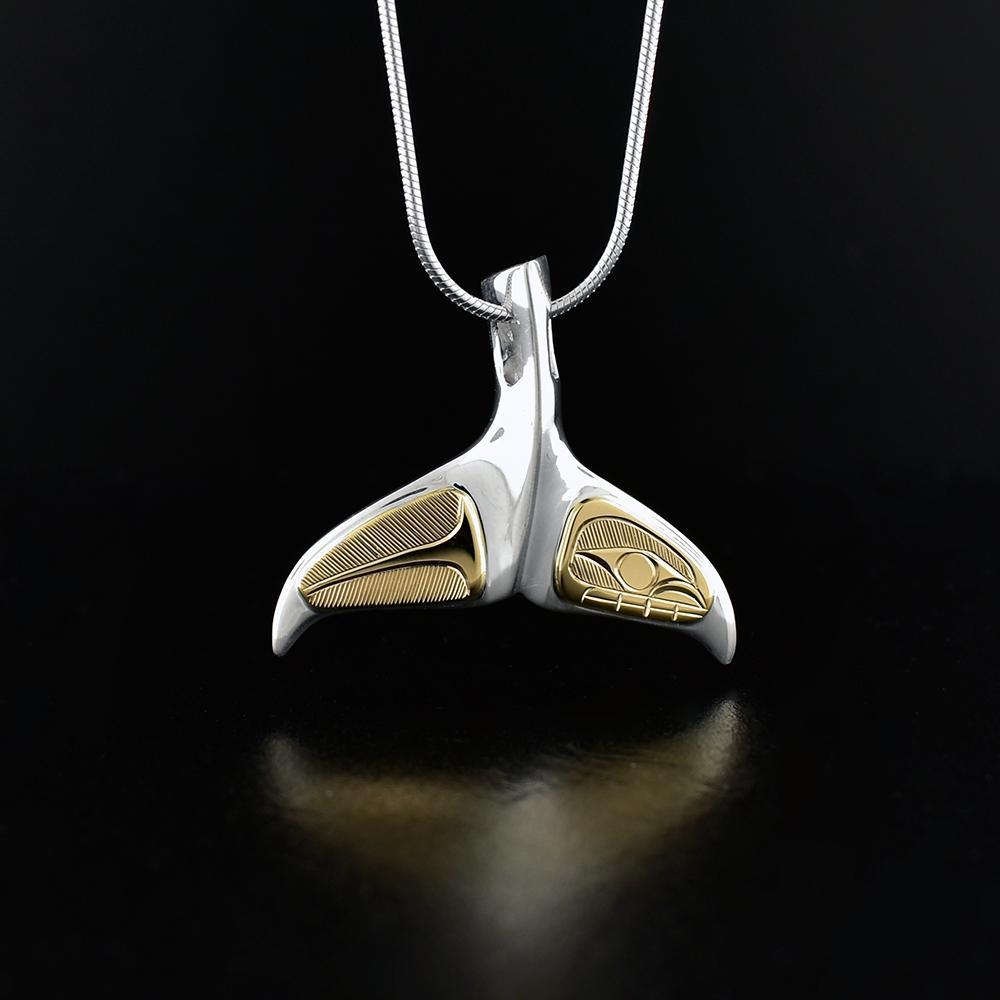 This gold and silver pendant is made from sterling silver and 14K gold and is shaped like the tail of the Orca. The fins of the tail are made from 14K gold and have carvings on the surface of them that depict the Orca.