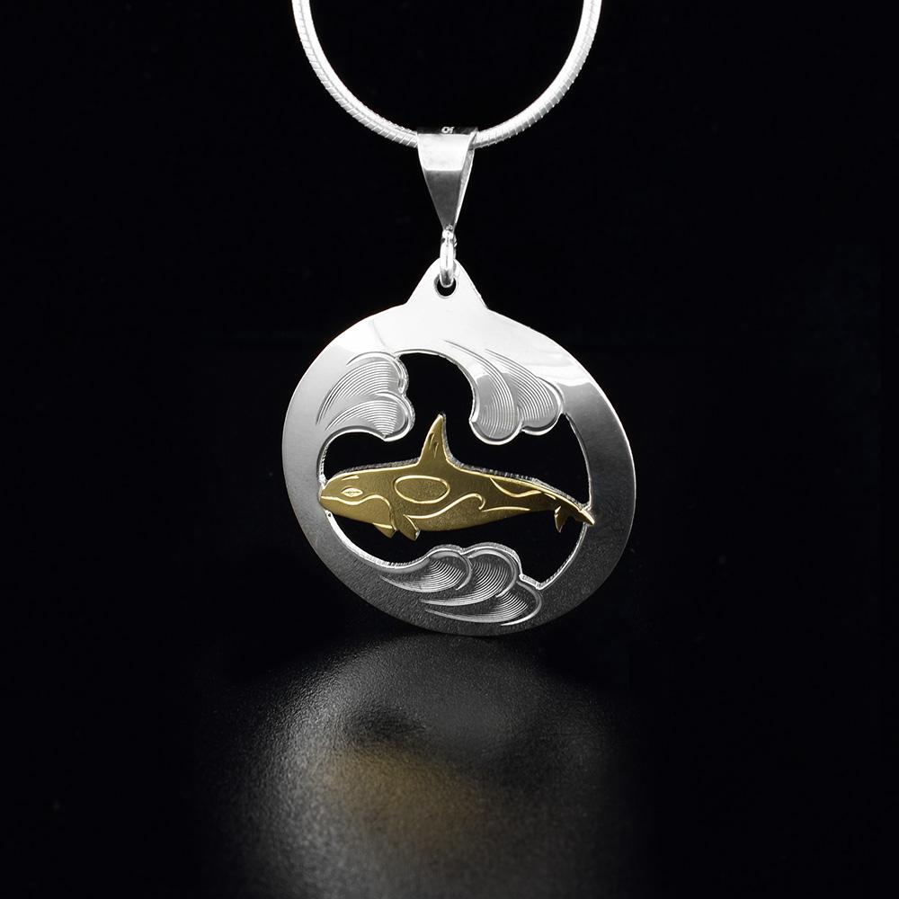 This Orca whale pendant is made form sterling silver and 10K gold. The piece has a sliver circle with a cutout in the middle; in it is a golden Orca. There are carvings and brushed patterns on the pendant depicting the Orca whale in the Ocean.