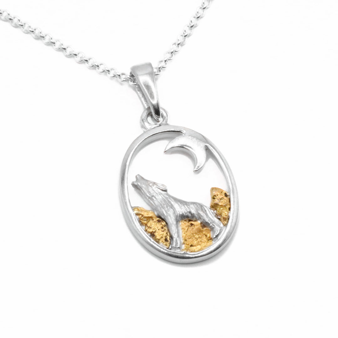 This 22K Gold Nugget Oval Wolf Pendant by Tom Gregorczyk. The pendant is an oval shape with an image of a wolf in the bottom half howling at a small moon in the top right corner. The artist has added 22k gold nuggets as the landscape behind the wolf.