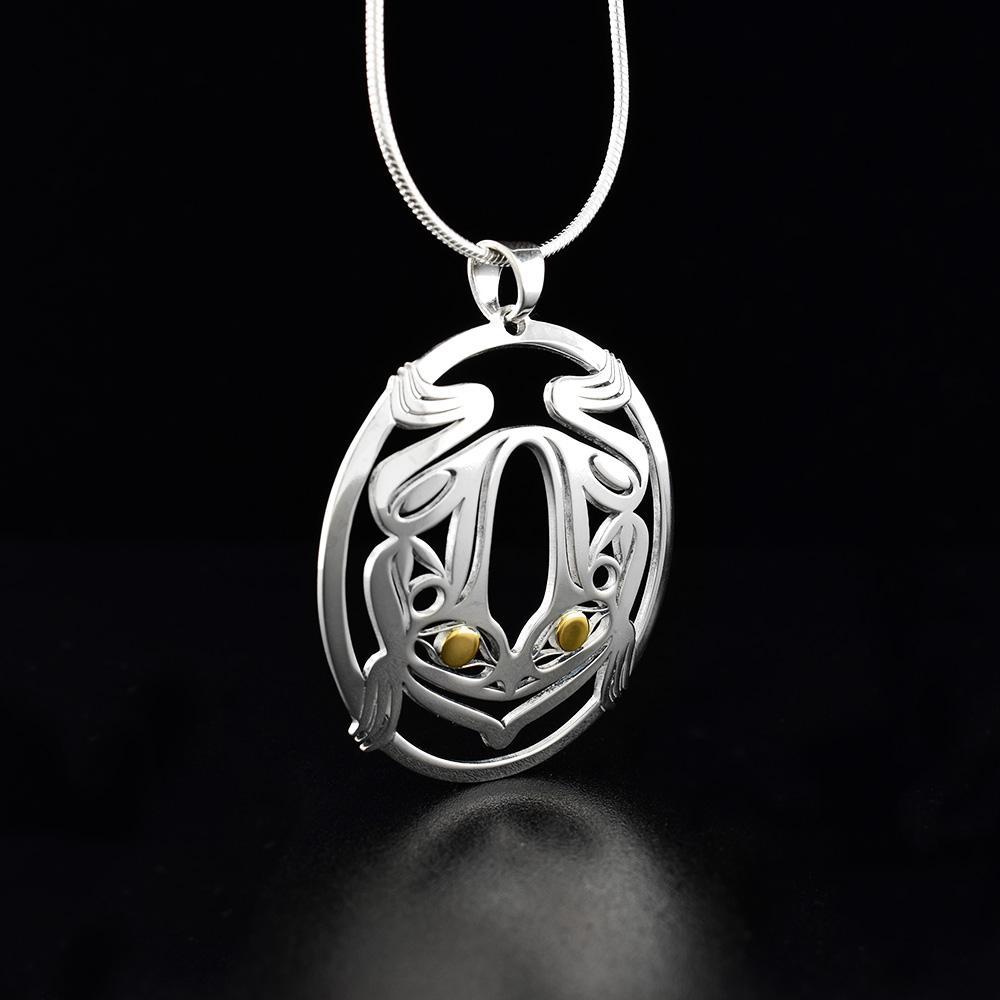 This silver and gold Frog pendant is made from sterling silver and 18K gold. The pendant is oval shaped and has cutout designs that depict the Frog. The eyes of the frog are made from 18K gold.