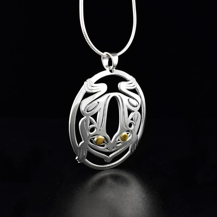 This silver and gold Frog pendant is made from sterling silver and 18K gold. The pendant is oval shaped and has cutout designs that depict the Frog. The eyes of the frog are made from 18K gold.