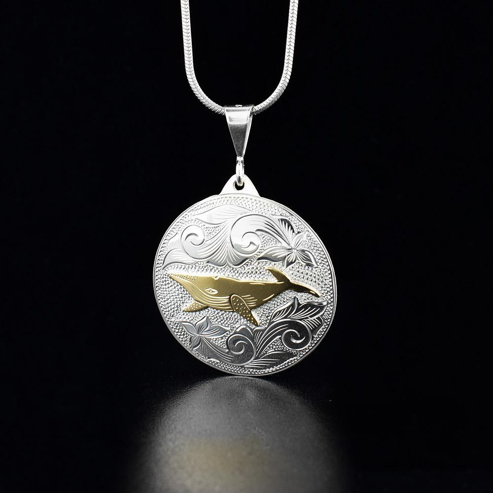 This whale pendant has been made from sterling silver and 10K gold with a circle shape. There are carvings on the surface of the pendant depicting a humpback whale in the ocean and the whale is done in gold.