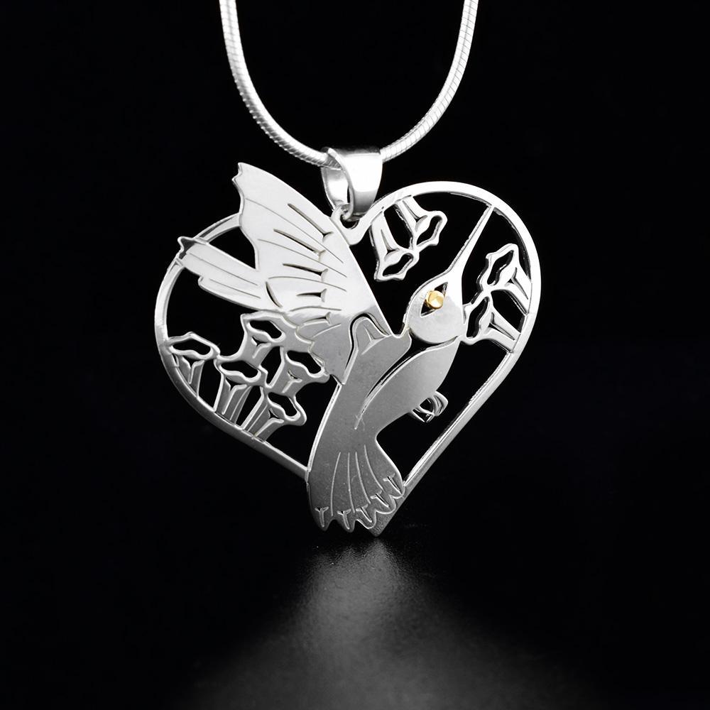 Heart Shaped Silver, Gold Hummingbird pendant by Grant Pauls. The pendant is in the shape of a heart. The artist has cut out the profile of a hummingbird's body mid-flight facing towards the right in the center of the pendant. Surrounding the hummingbird are flowers. The artist has put a 18k gold accent in the hummingbird's eye.
