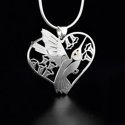Sterling Silver and 18K Gold Hummingbird Heart Pendant by Grant Pauls. The pendant is in the shape of a heart. The artist has cut out the profile of a hummingbird's body mid-flight facing towards the right in the center of the pendant. Surrounding the hummingbird are flowers. The artist has put a 18k gold accent in the hummingbird's eye.